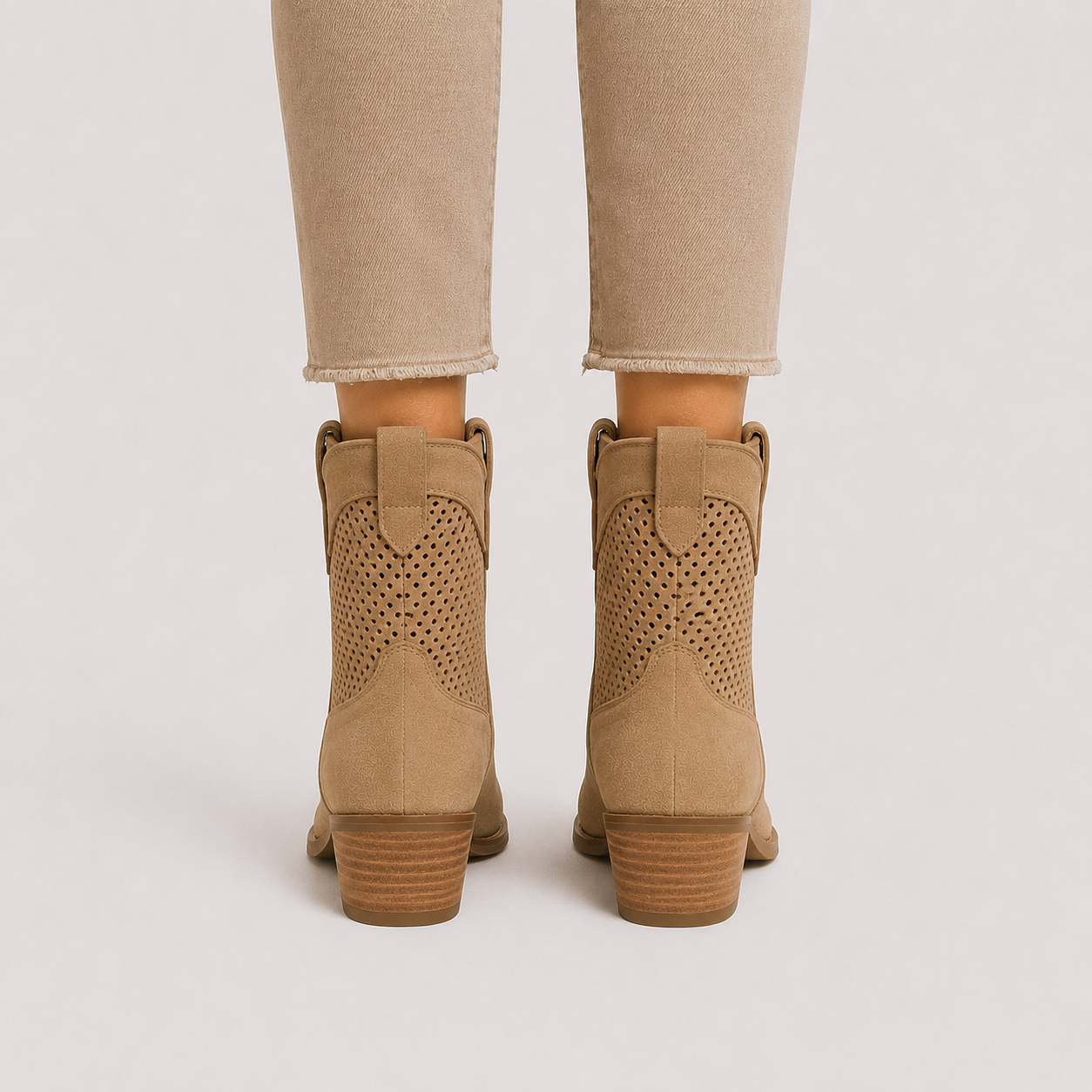 A pair of beige suede ankle boots with perforated side panels and stacked block heels, shown from the back on a model wearing beige pants against a light gray background.