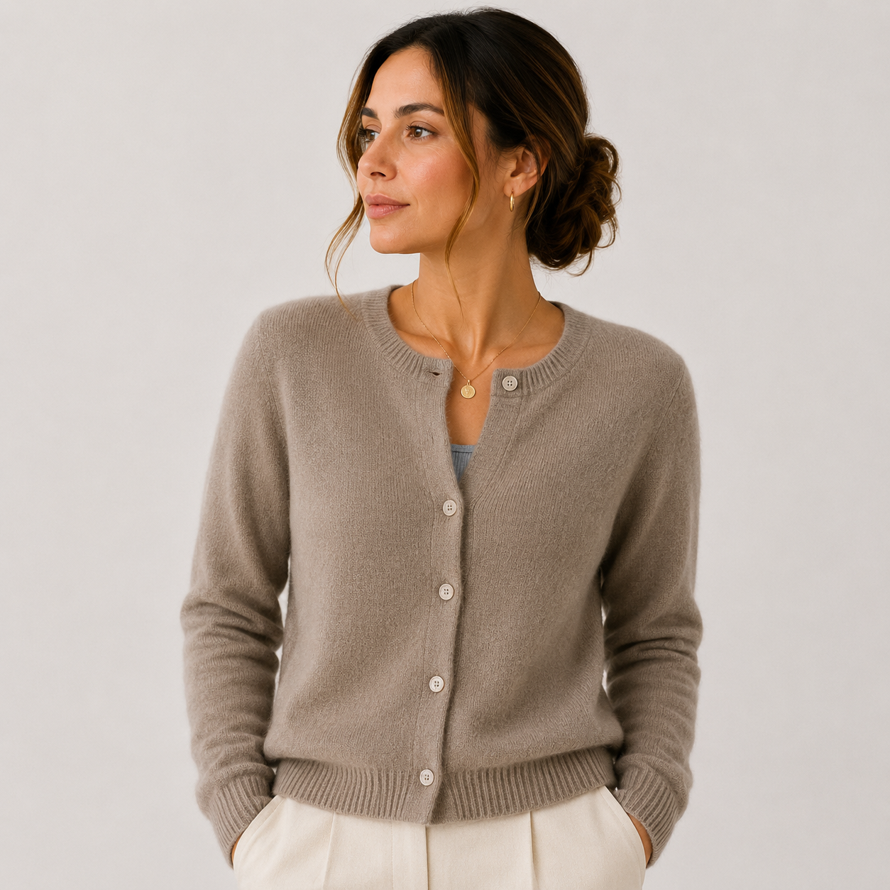 A woman wearing a taupe knit cardigan sweater with white buttons, styled with cream trousers against a light gray studio background.