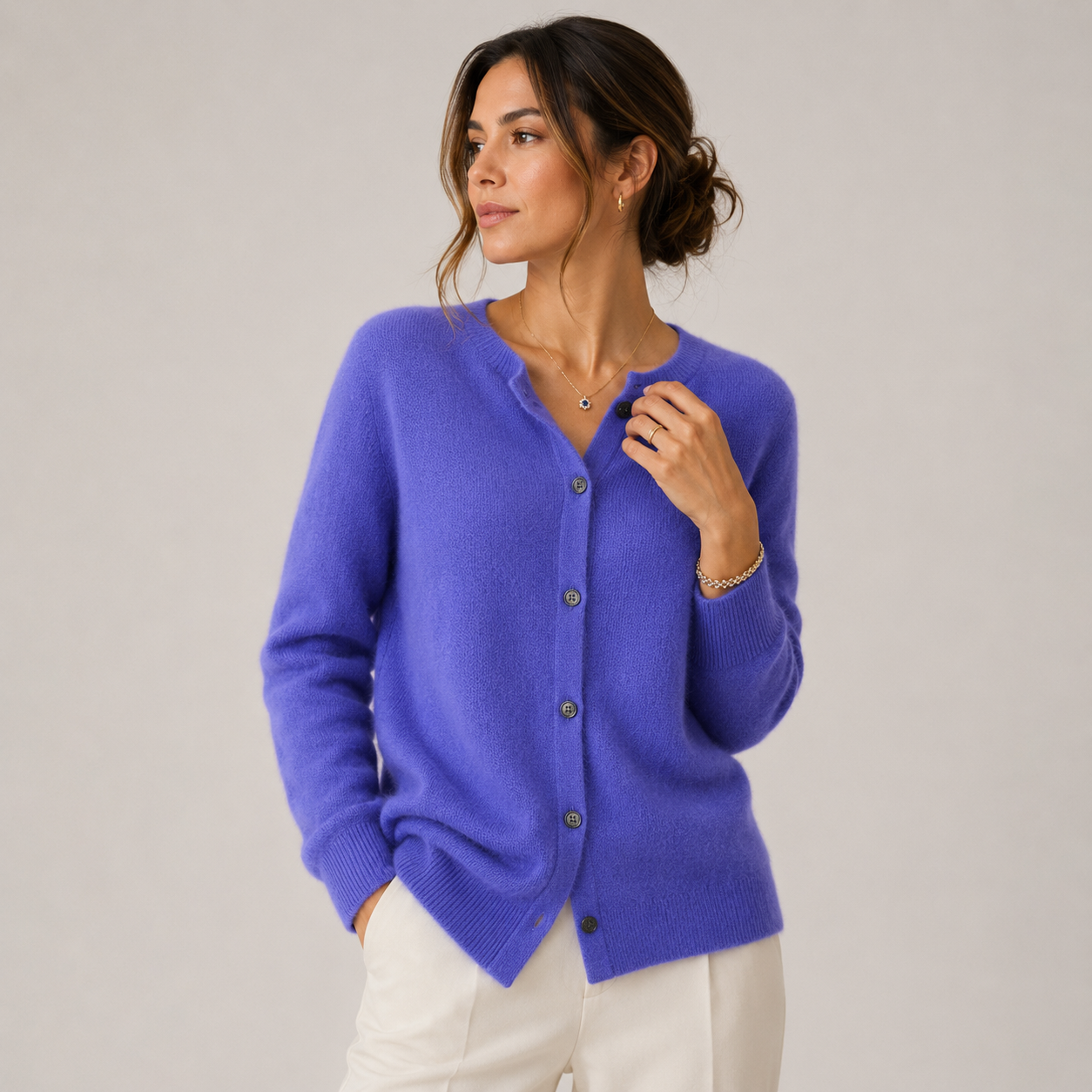 A woman wearing a vibrant periwinkle blue knit cardigan sweater with dark buttons, paired with white pants, photographed against a neutral light gray studio background.