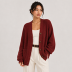 A woman models a ribbed knit cardigan in deep burgundy red, worn open over a white tank top and cream pants with a brown belt, against a light gray studio background.