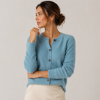 A woman wearing a light blue knit cardigan sweater with dark buttons, paired with cream-colored pants, photographed against a neutral light gray studio background.