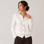 A woman wearing a cream-colored knit cardigan sweater with dark buttons, paired with dark brown trousers, photographed against a neutral light gray studio background.