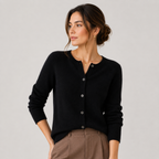 A woman wearing a black long-sleeve button-front cardigan sweater and brown trousers, photographed against a plain light gray studio background.