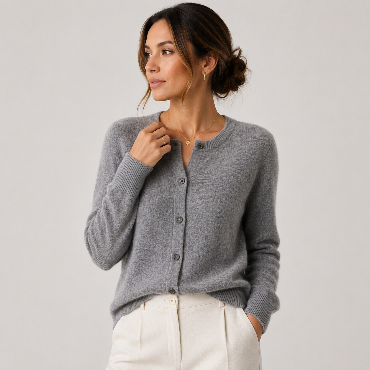 A woman wearing a light gray knit cardigan with dark buttons and white pants, posing against a light gray studio background.