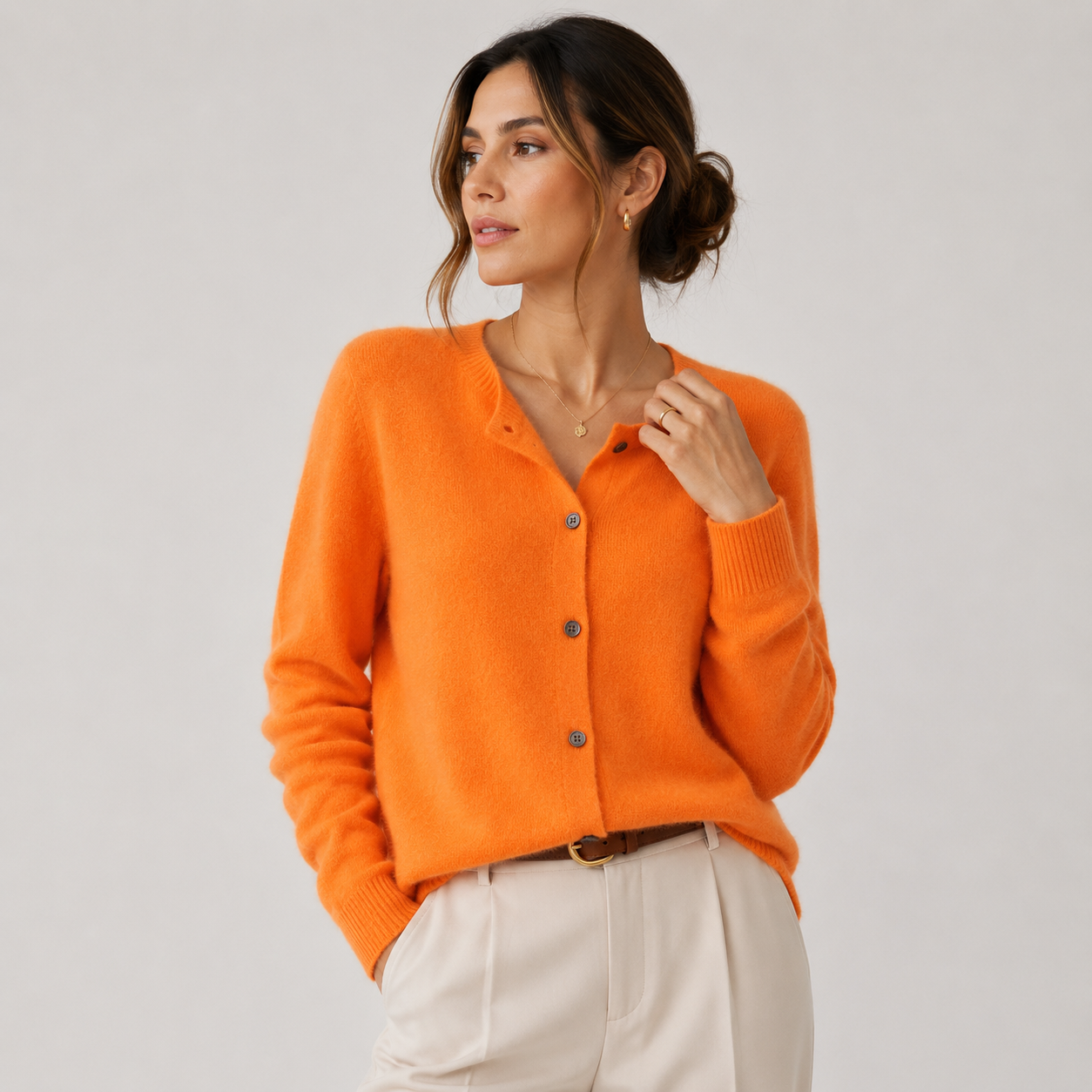 A woman wearing a bright orange knit cardigan sweater with brown buttons, paired with cream-colored trousers, photographed against a neutral light gray studio background.