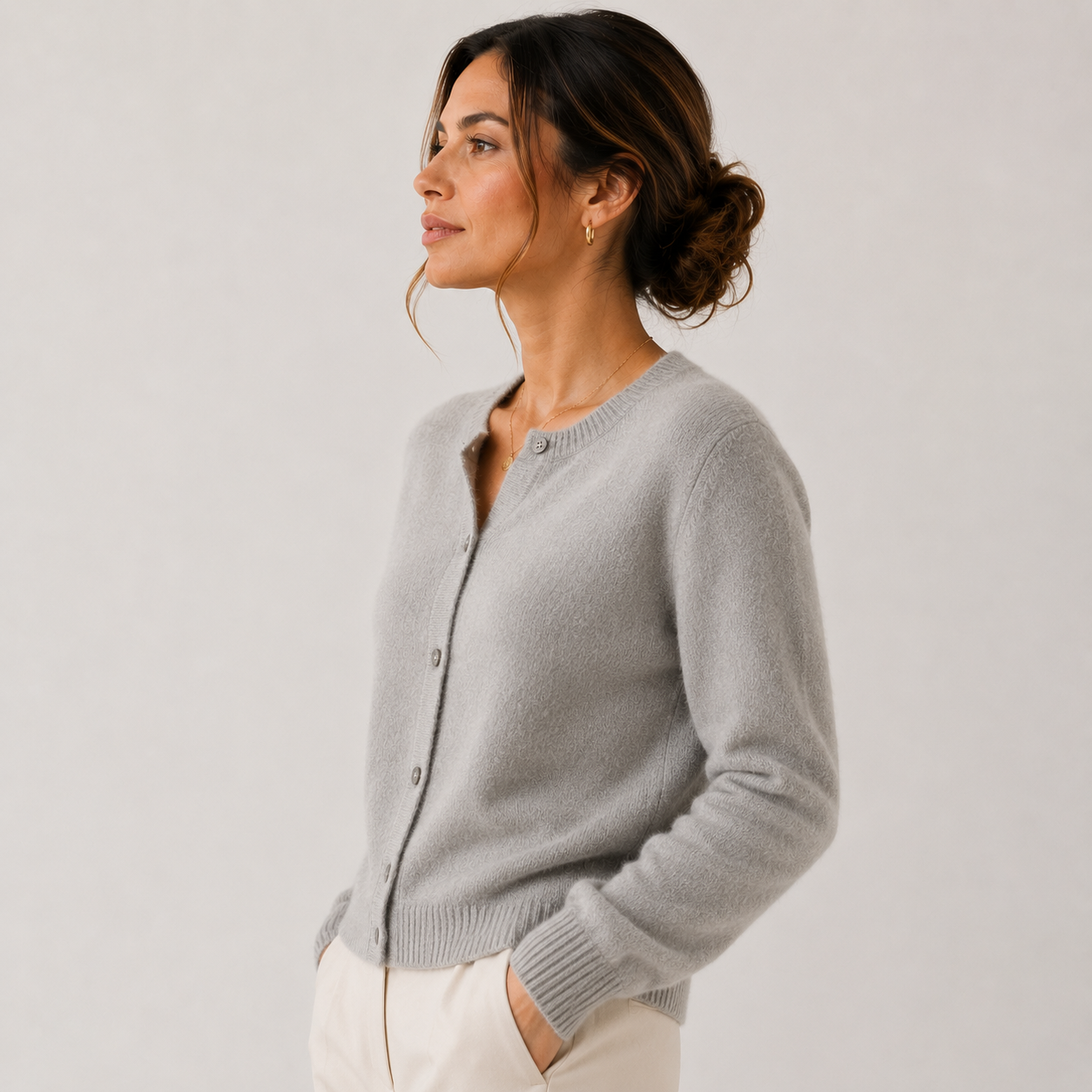 A woman wearing a light gray knit cardigan with a button-front closure and ribbed collar, cuffs, and hem, paired with off-white pants, photographed against a plain light gray studio background.