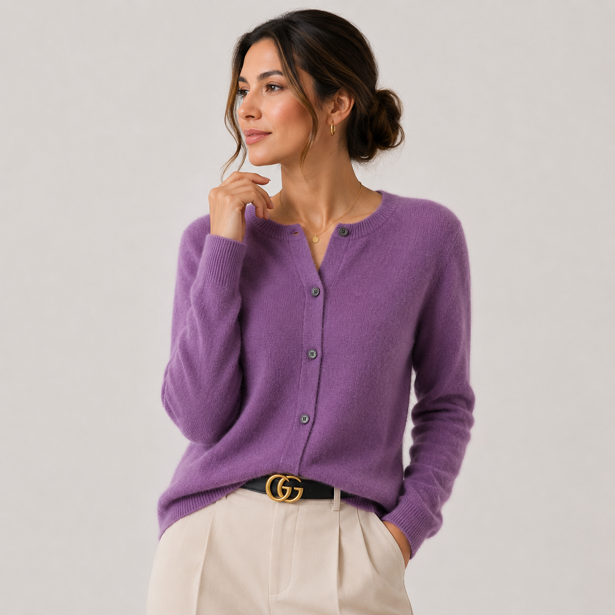 A woman wearing a purple knit cardigan sweater with buttons, paired with cream trousers and a black belt with a gold buckle, photographed against a light gray studio background.