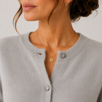 Close-up of a woman wearing a light gray ribbed knit cardigan with silver buttons and a delicate gold necklace with a round pendant, against a plain light background.