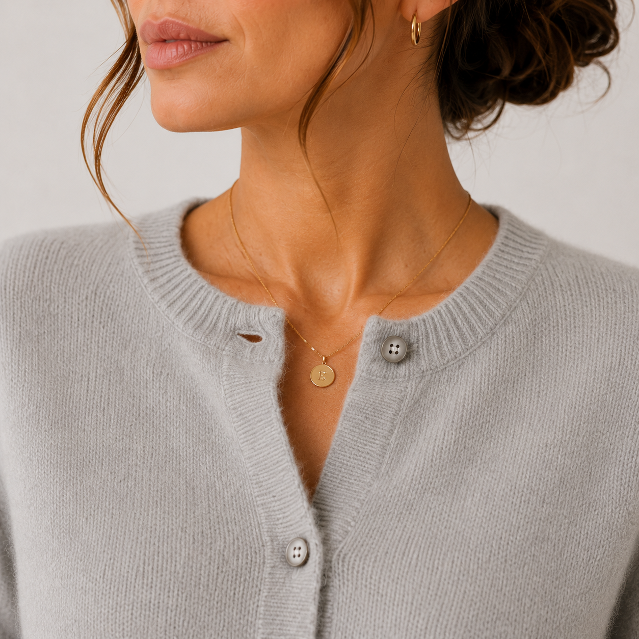 Close-up of a woman wearing a light gray ribbed knit cardigan with silver buttons and a delicate gold necklace with a round pendant, against a plain light background.