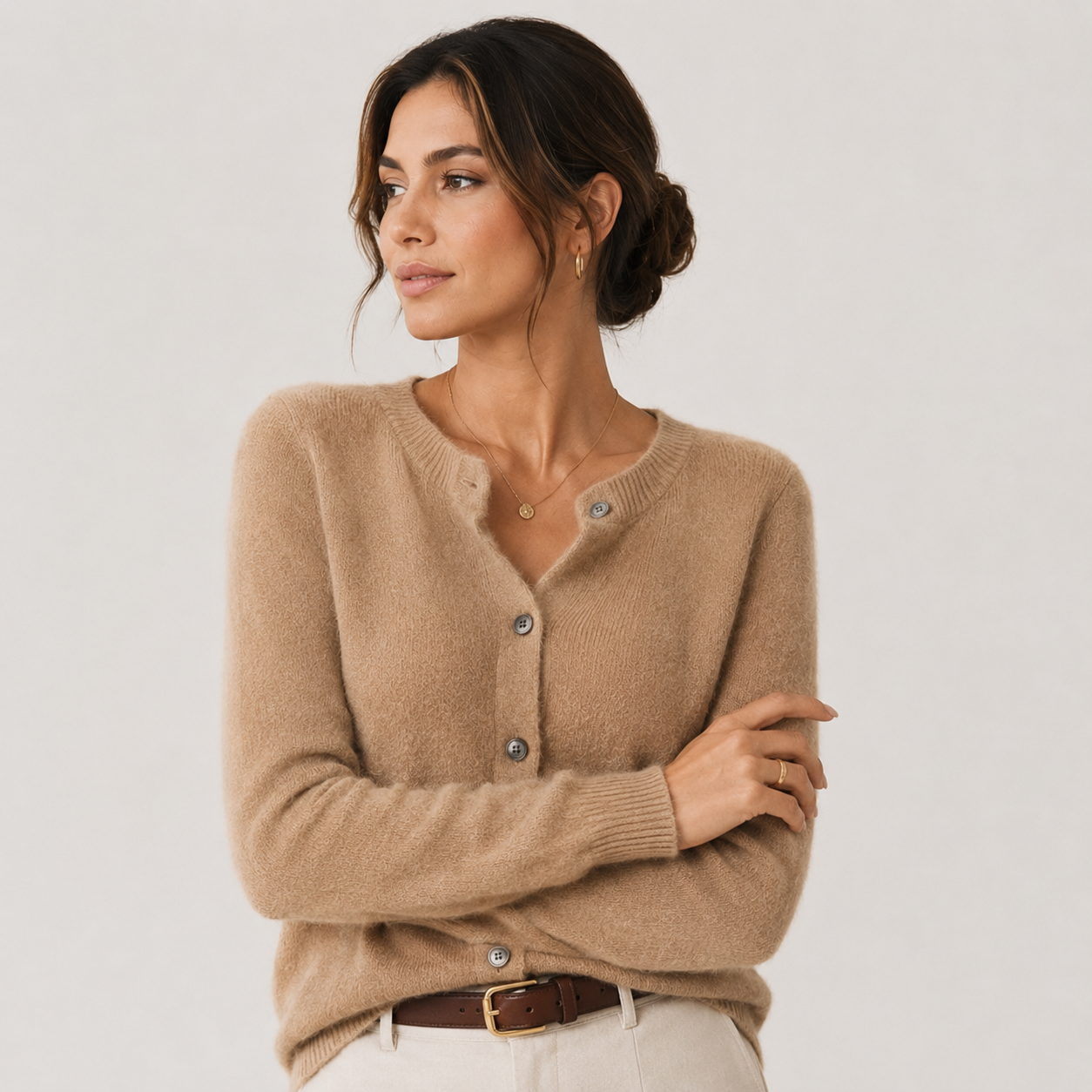 A woman wearing a soft camel beige knit cardigan sweater with a split neckline and button-front closure, paired with light pants and a brown belt, photographed against a plain light gray studio background.
