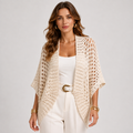 A woman models a cream-colored, open-knit crochet cardigan over a white tank top and white trousers with a gold belt buckle, set against a plain light gray studio background.