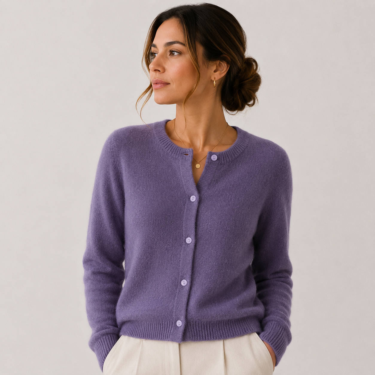 A woman wearing a lavender purple knit cardigan sweater with white buttons, paired with cream-colored pants, photographed against a neutral light gray studio background.