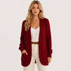 A woman modeling a chunky knit cardigan in deep burgundy red, worn over a white camisole and cream pants with a brown belt, against a plain white studio background.