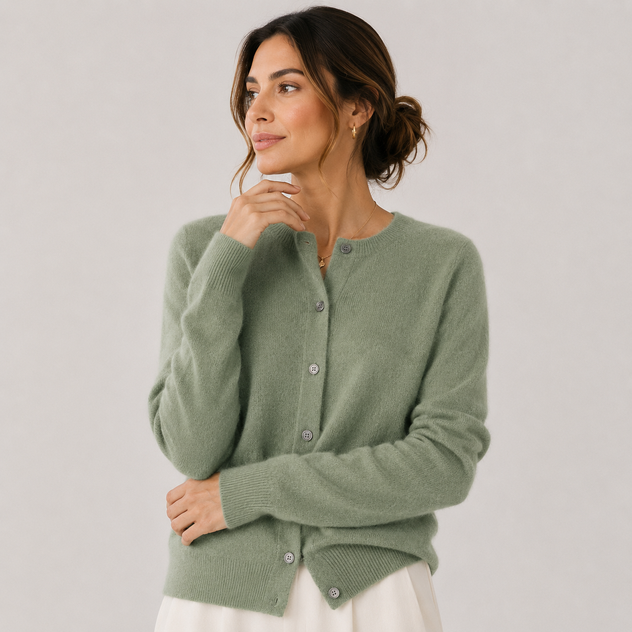 A woman wearing a sage green knit cardigan sweater with buttons, paired with a white skirt, photographed against a neutral light gray studio background.