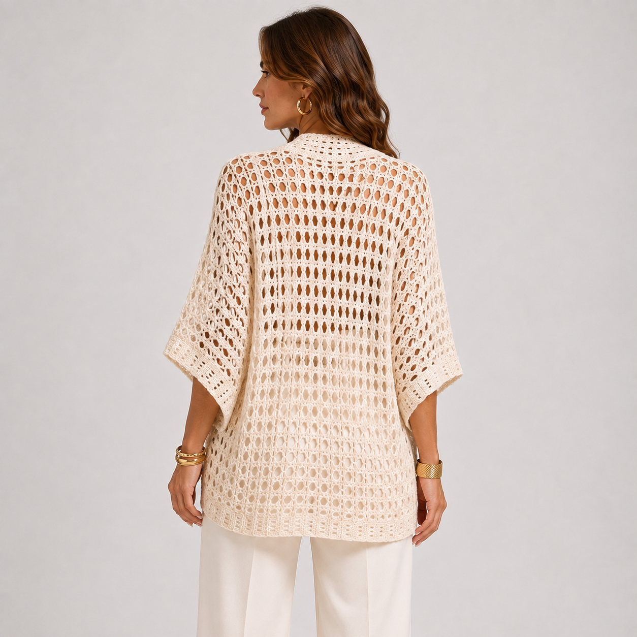 A woman shown from the back wearing a cream-colored, open-knit crochet cardigan with wide three-quarter sleeves, paired with white pants, against a light gray studio background.