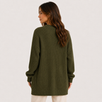 A woman shown from the back wearing an oversized olive green ribbed knit sweater, paired with off-white pants, against a plain light gray studio background.