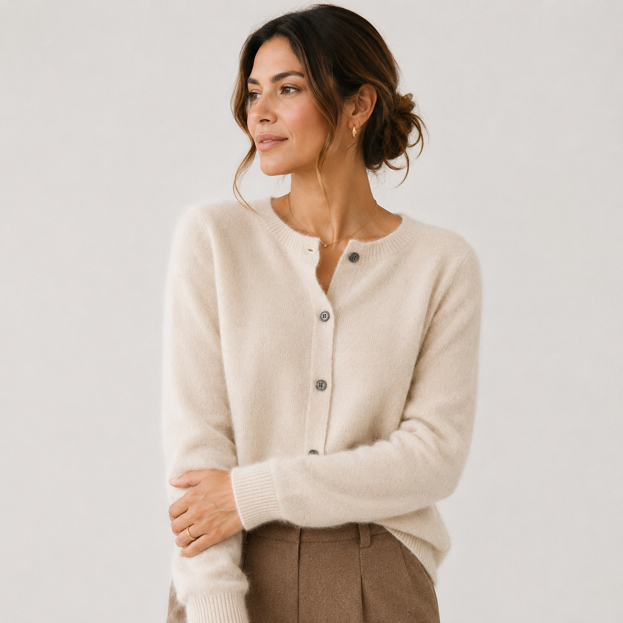 A woman wearing a cream-colored, long-sleeve knit cardigan with dark buttons, paired with brown trousers, photographed against a solid light gray background.