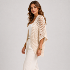 A woman models a cream open-knit crochet cardigan over a white tank top and white pants, accessorized with gold bracelets and earrings, against a light gray studio background.