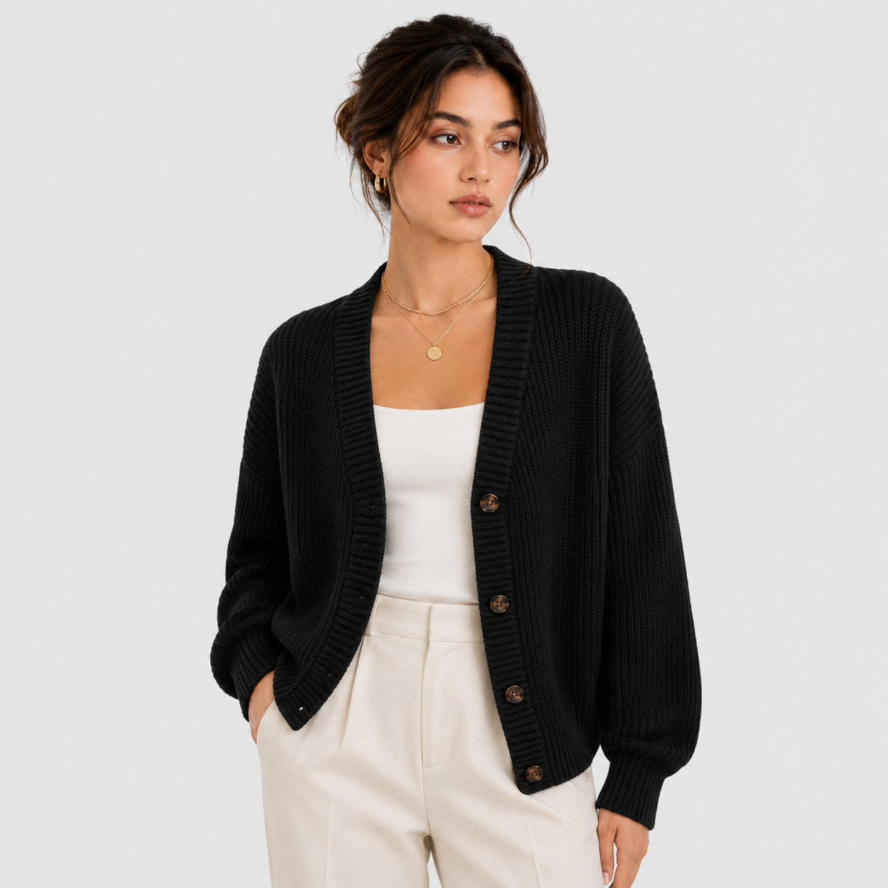 A woman wearing a black ribbed knit cardigan with brown buttons, styled over a white tank top and cream trousers, photographed against a light gray studio background.