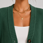 A close-up of a person wearing layered gold necklaces, including a chunky chain necklace and a delicate chain with a small round disc pendant, styled over a white top and green cardigan.