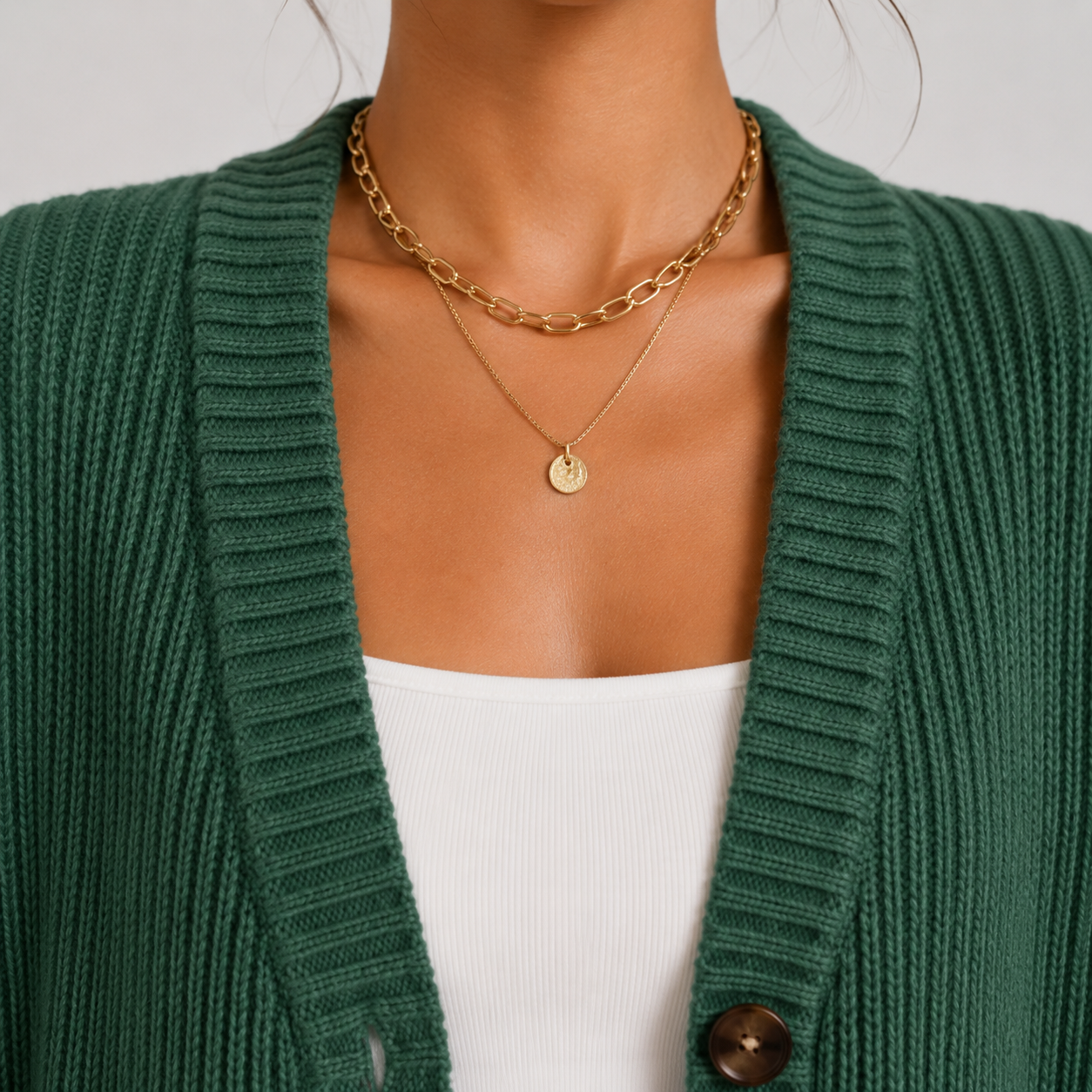 A close-up of a person wearing layered gold necklaces, including a chunky chain necklace and a delicate chain with a small round disc pendant, styled over a white top and green cardigan.