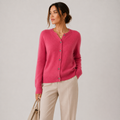 A woman wearing a bright pink knit cardigan with buttons and light beige trousers, holding a beige handbag against a plain light gray studio background.