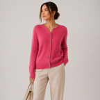A woman wearing a bright pink knit cardigan with buttons and light beige trousers, holding a beige handbag against a plain light gray studio background.
