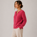 A woman wearing a bright pink knit cardigan sweater with dark buttons and cream pants, standing against a light gray studio background.