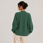 A woman shown from the back wearing an oversized ribbed knit sweater in a muted green color, paired with light beige trousers, against a plain light gray background.