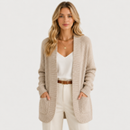 A woman modeling a long, open-front ribbed knit cardigan in a light oatmeal beige color, worn over a white camisole and cream pants with a brown belt, against a plain white studio background.