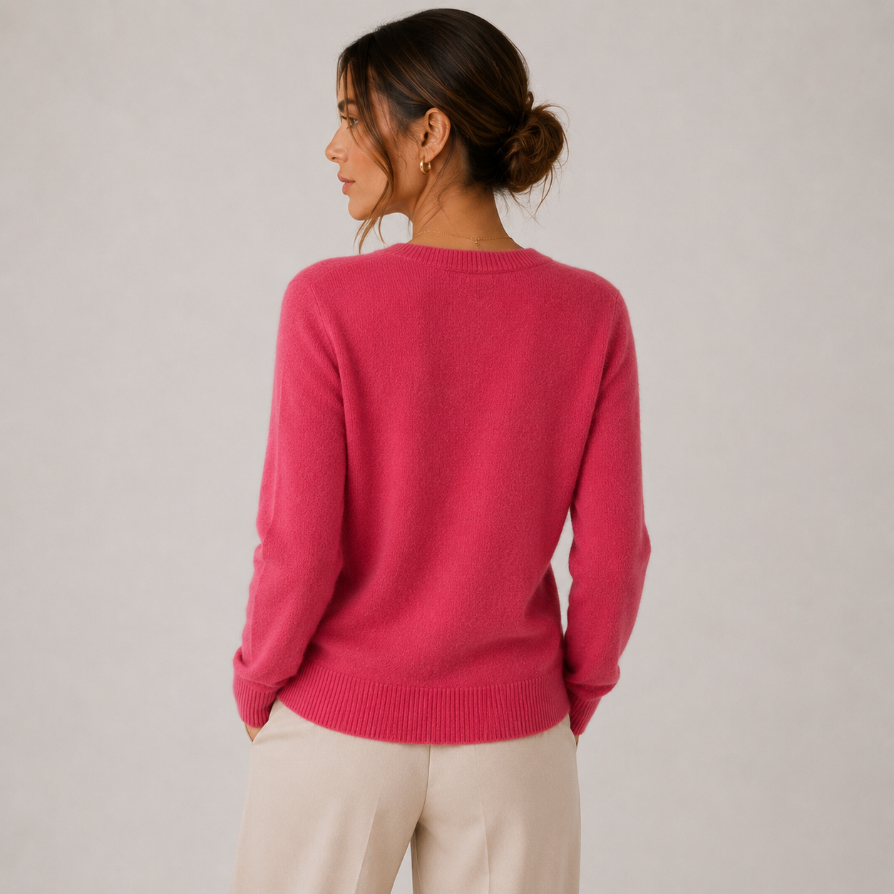 A woman shown from the back wearing a bright pink knit sweater and light beige trousers, standing against a plain light gray studio background.