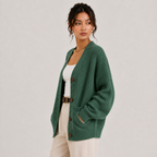 A woman models a green ribbed open-front cardigan with brown buttons, worn over a white tank top and cream pants, against a plain light gray studio background.