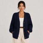 A woman models a navy blue ribbed knit cardigan with brown buttons, worn over a white tank top and cream pants, against a light gray studio background.