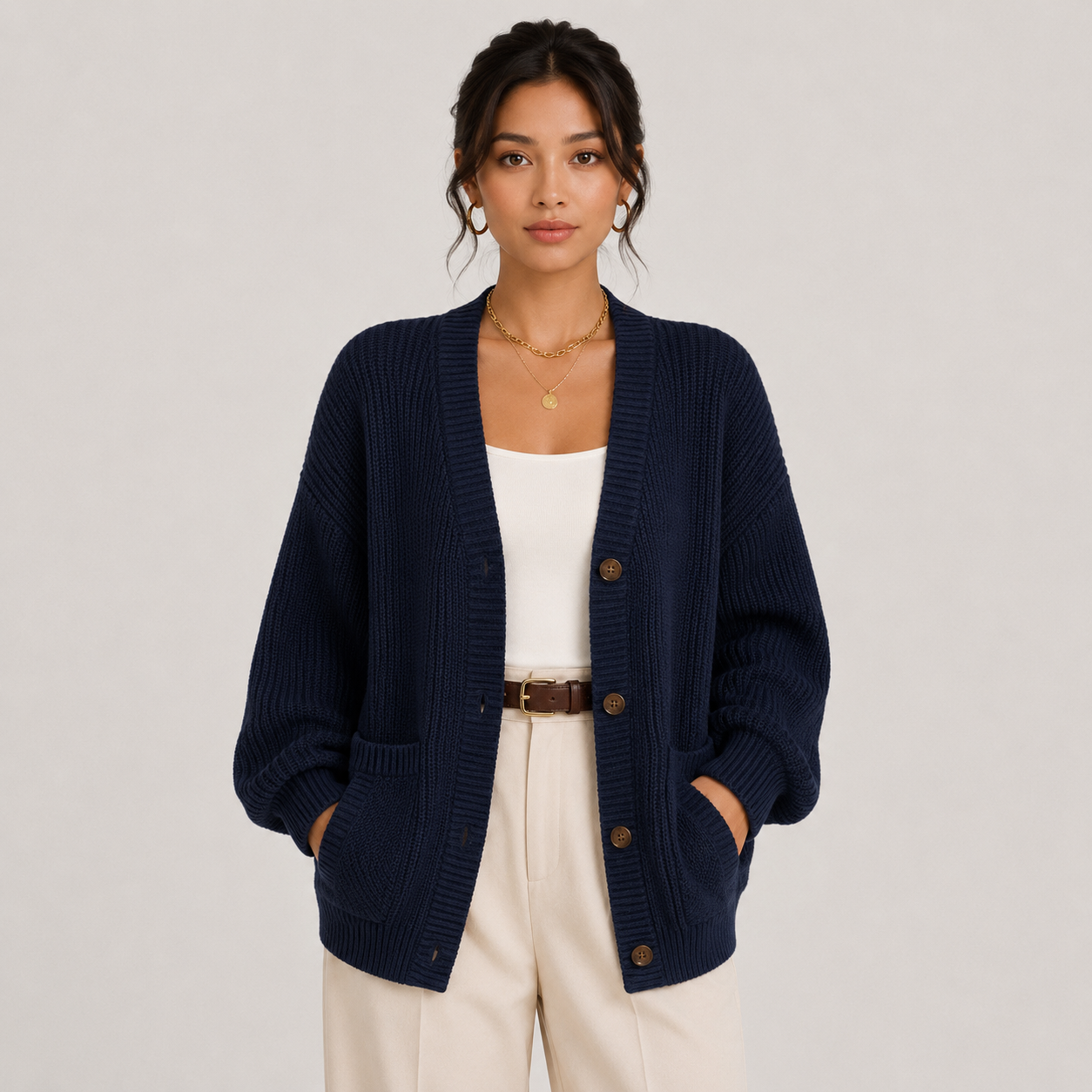 A woman models a navy blue ribbed knit cardigan with brown buttons, worn over a white tank top and cream pants, against a light gray studio background.