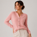 A woman wearing a light pink fuzzy button-up cardigan sweater and cream trousers, posing against a neutral light gray background.