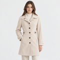 A woman modeling a light beige mid-length coat with a notched lapel, dark brown buttons, and a matching fabric belt, paired with a white turtleneck and beige trousers against a plain white studio background.