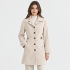 A woman modeling a light beige mid-length coat with a notched lapel, dark brown buttons, and a matching fabric belt, paired with a white turtleneck and beige trousers against a plain white studio background.