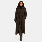 A woman modeling a long dark brown wool-blend coat with a matching waist tie belt, worn over a black turtleneck and brown knee-high boots, against a light gray studio background.