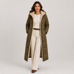 A woman models a long olive green puffer coat with a beige sherpa-lined hood and interior, styled with a cream top, cream wide-leg trousers, and brown shoes against a light gray studio background.