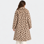 A woman viewed from behind wearing a beige faux-fur coat with a dark brown floral pattern, paired with white pants, against a plain light gray background.
