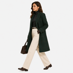 A woman modeling a dark green windowpane plaid coat over a black turtleneck, cream wide-leg trousers, and dark brown loafers, carrying a brown leather handbag against a light gray studio background.
