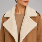 Close-up of a model wearing a camel brown shearling-lined coat over a beige knit sweater, against a neutral gray background.