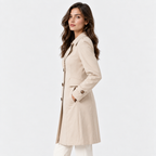 A woman wearing a light beige trench coat with white pants, shown from a side profile against a plain white studio background.