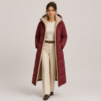 A woman models a long burgundy puffer coat with a cream sherpa-lined hood and interior, worn over a cream sweater and matching trousers, against a light gray studio background.