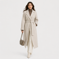 A woman models a long, light beige belted coat over a brown turtleneck, matching wide-leg trousers, and beige ankle boots, while holding a taupe handbag against a plain light gray studio background.
