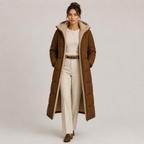 A woman models a long brown puffer coat with a beige sherpa-lined hood and interior, styled with a cream sweater, cream trousers, and brown shoes against a light gray studio background.