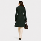 A woman shown from the back wearing a dark green windowpane plaid coat, cream trousers, black gloves, and carrying a brown leather handbag against a light gray studio background.