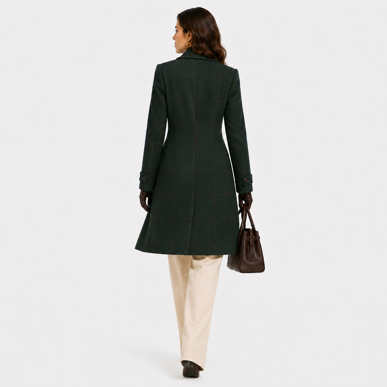 A woman shown from the back wearing a dark green windowpane plaid coat, cream trousers, black gloves, and carrying a brown leather handbag against a light gray studio background.