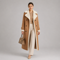A woman models a long camel-colored double-breasted coat with a cream shearling-style collar and lining, paired with cream trousers, a beige top, and a structured taupe handbag against a neutral gray studio background.