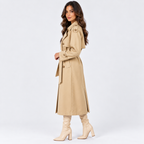 A woman modeling a mid-length beige trench coat with a matching belt, paired with beige heeled boots, against a plain light gray studio background.